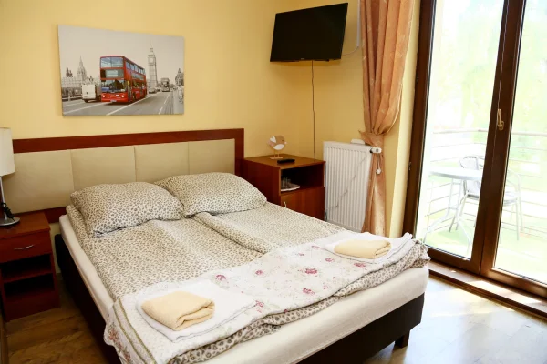 Sypialnia z dużym podwójnym łóżkiem nakrytym beżową narzutą i kwiecistą kapą, złożone ręczniki, telewizor i balkon.