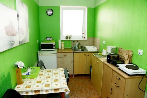 Aneks kuchenny w pokoju z oknem — zielone ściany, kuchenka, mikrofalówka, lodówka i stół z obrusem w kratę.