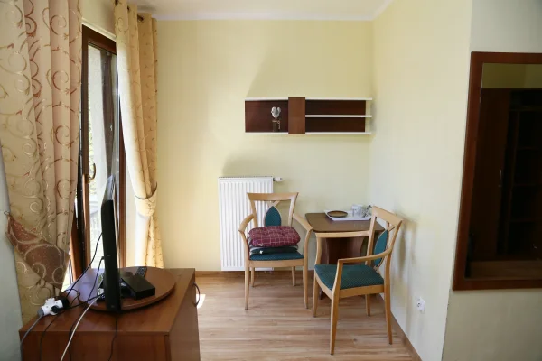 W pokoju dwa krzesła na zielonych siedziskach, stolik z tacą, telewizor na drewnianej komodzie, balkon z beżowymi zasłonami.