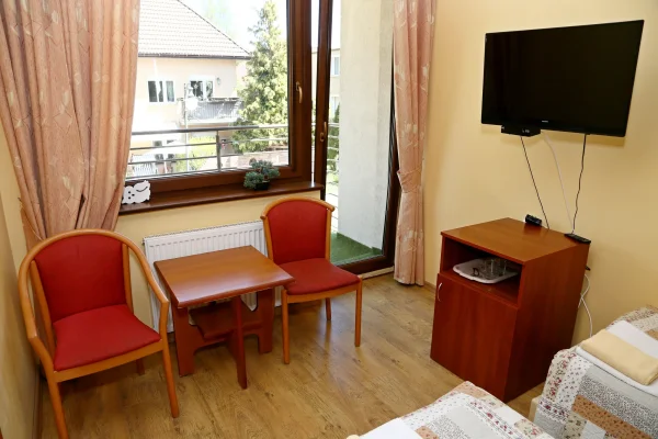 Dwa czerwone fotele przy drewnianym stoliku, telewizor na ścianie i wyjście na balkon.