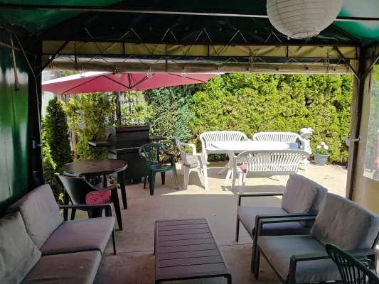 Zadaszona altana w ogrodzie pensjonatu w Pobierowie — biały stolik z krzesłami, czerwony parasol, grill i szare fotele.
