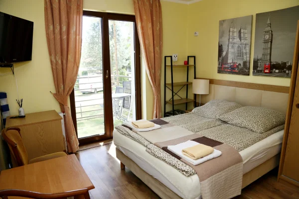 Pokój z szerokim łóżkiem w panterkę, balkon z białym stolikiem, żółte ściany i dwa czarno-białe obrazy Londynu.