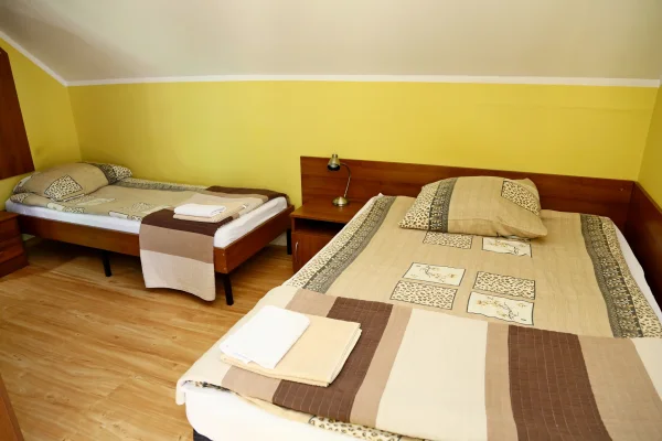 Pokój z żółtymi ścianami — podwójne łóżko z patchworkową narzutą, drugie pojedyncze, ręczniki i mosiężna lampka na szafce.